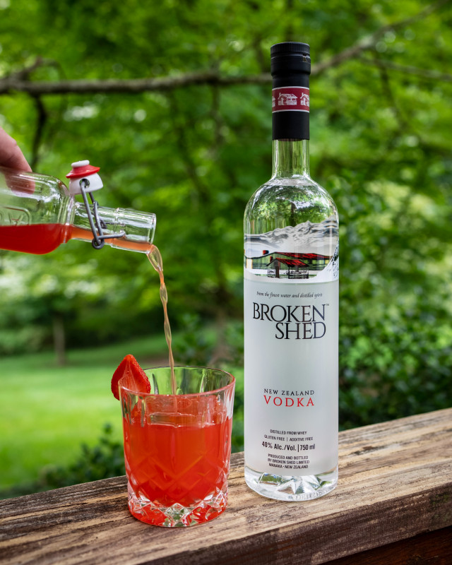 A bright red cocktail poured into a glass next to a bottle of Broken Shed